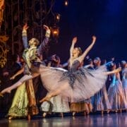 Ballet dancers perform on stage in elaborate costumes, with two lead dancers leaping gracefully at the front