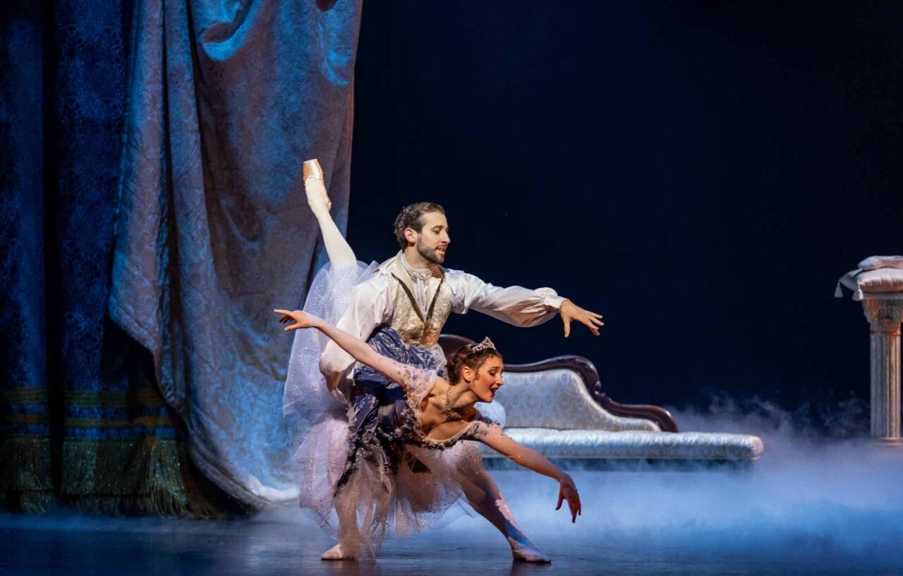 A male and female ballet dancer perform on stage in elaborate costumes. The man supports the woman in an arabesque pose, with ornate drapery, a classical sofa, and stage fog in the background.