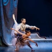 A male and female ballet dancer perform on stage in elaborate costumes. The man supports the woman in an arabesque pose, with ornate drapery, a classical sofa, and stage fog in the background.