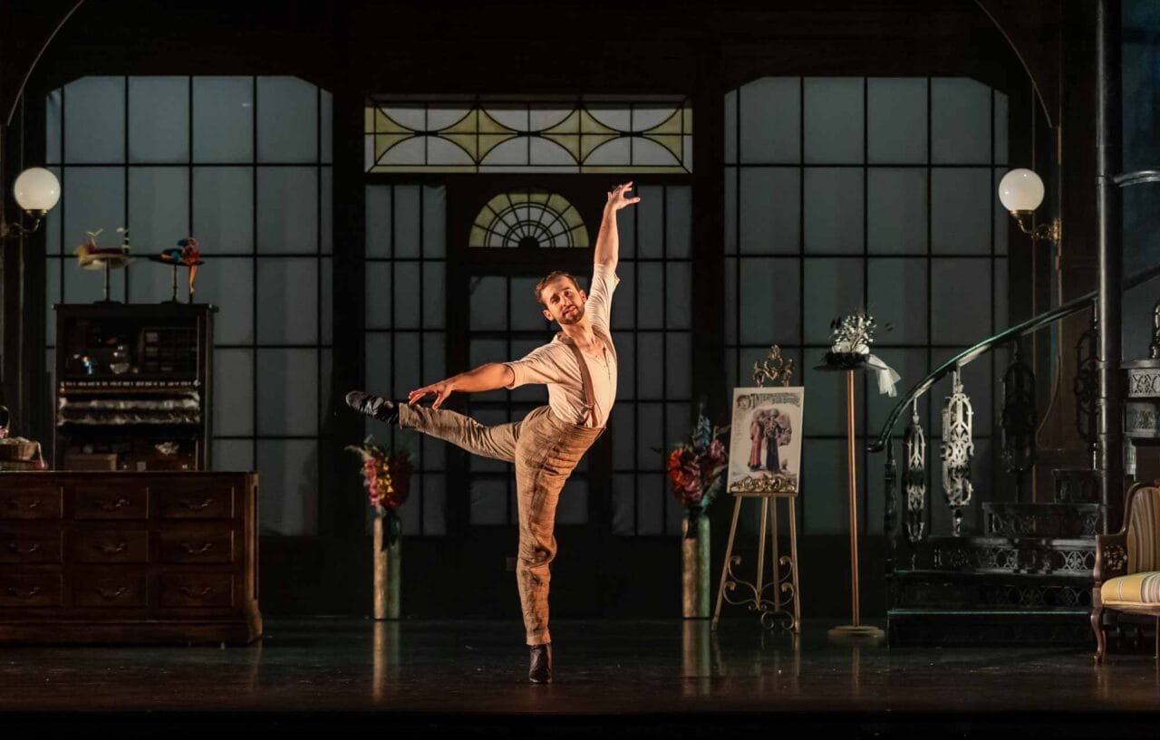 A ballet dancer in beige costume performs an arabesque on stage, one arm raised.