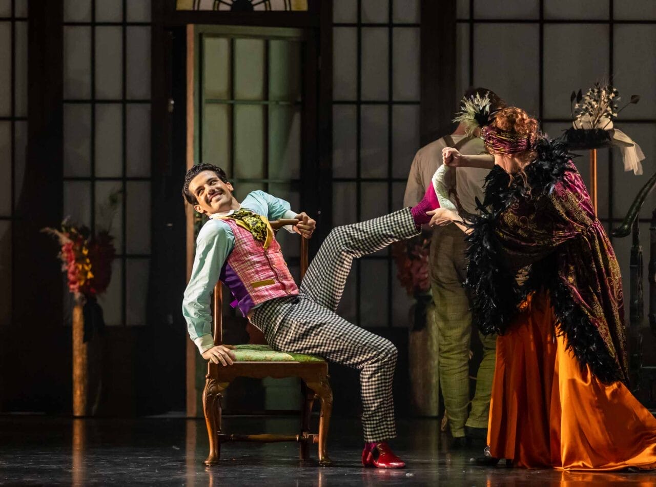A man in colourful clothing sits on a chair, smiling and lifting his leg, while a woman in an orange and black outfit helps him put on or remove a shoe.