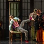 A man in colourful clothing sits on a chair, smiling and lifting his leg, while a woman in an orange and black outfit helps him put on or remove a shoe.