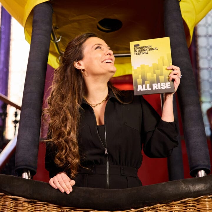 A woman in a black outfit stands in a hot air balloon basket, smiling and holding up an "Edinburgh International Festival: All Rise" booklet, with Capital Theatres’ grand stairs and large windows in the background.