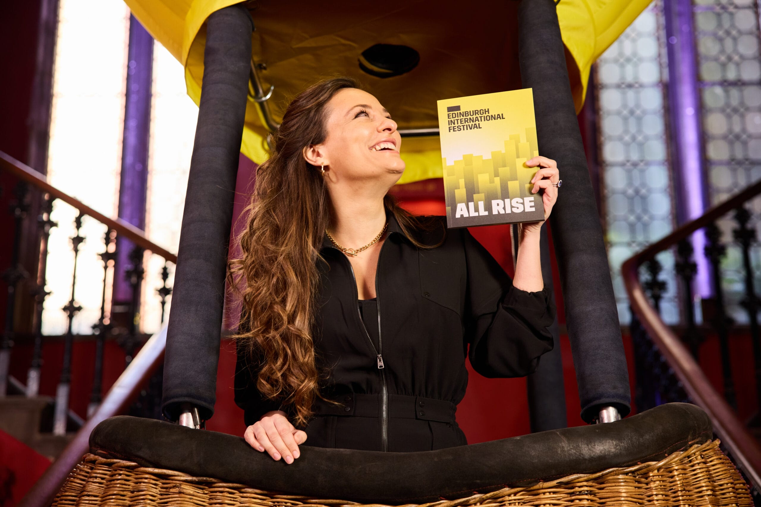 A woman in a black outfit stands in a hot air balloon basket, smiling and holding up an "Edinburgh International Festival: All Rise" booklet, with Capital Theatres’ grand stairs and large windows in the background.