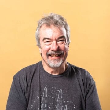 A man with grey hair and a beard is smiling, wearing a dark graphic T-shirt with spaceships on it, in front of a plain yellow background.