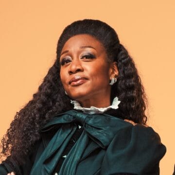 A woman with long curly hair, wears a dark green outfit with a large bow at the neck as she poses confidently