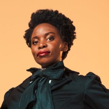A woman with natural short curly hair and red lipstick wears a dark blouse with a large bow at the neck, She looks confidently at the camera.