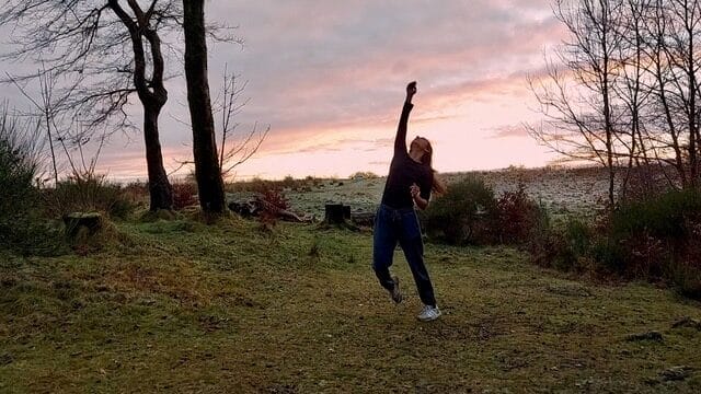 A person stands on a grassy area near trees, reaching one arm upward with the other arm down.