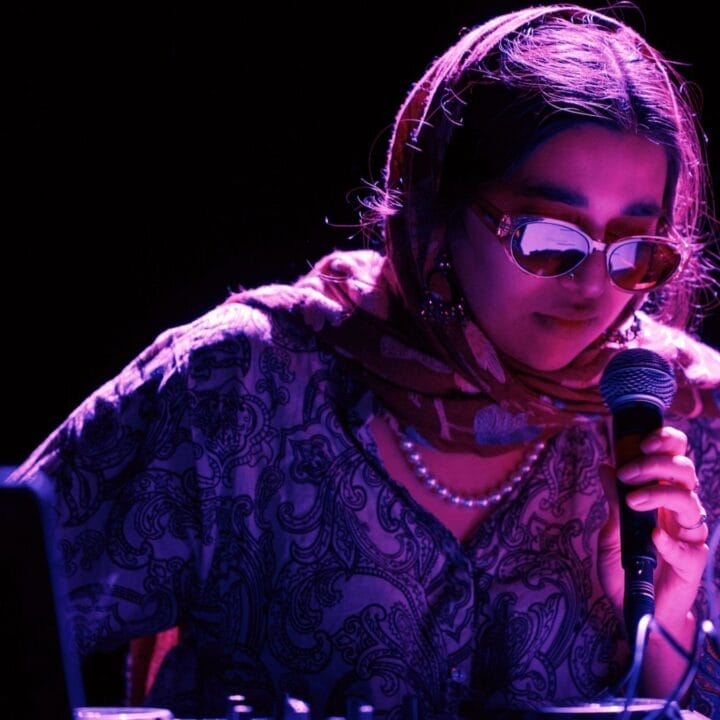 A person wearing sunglasses and a patterned scarf holds a microphone whilst operating audio equipment under purple lighting