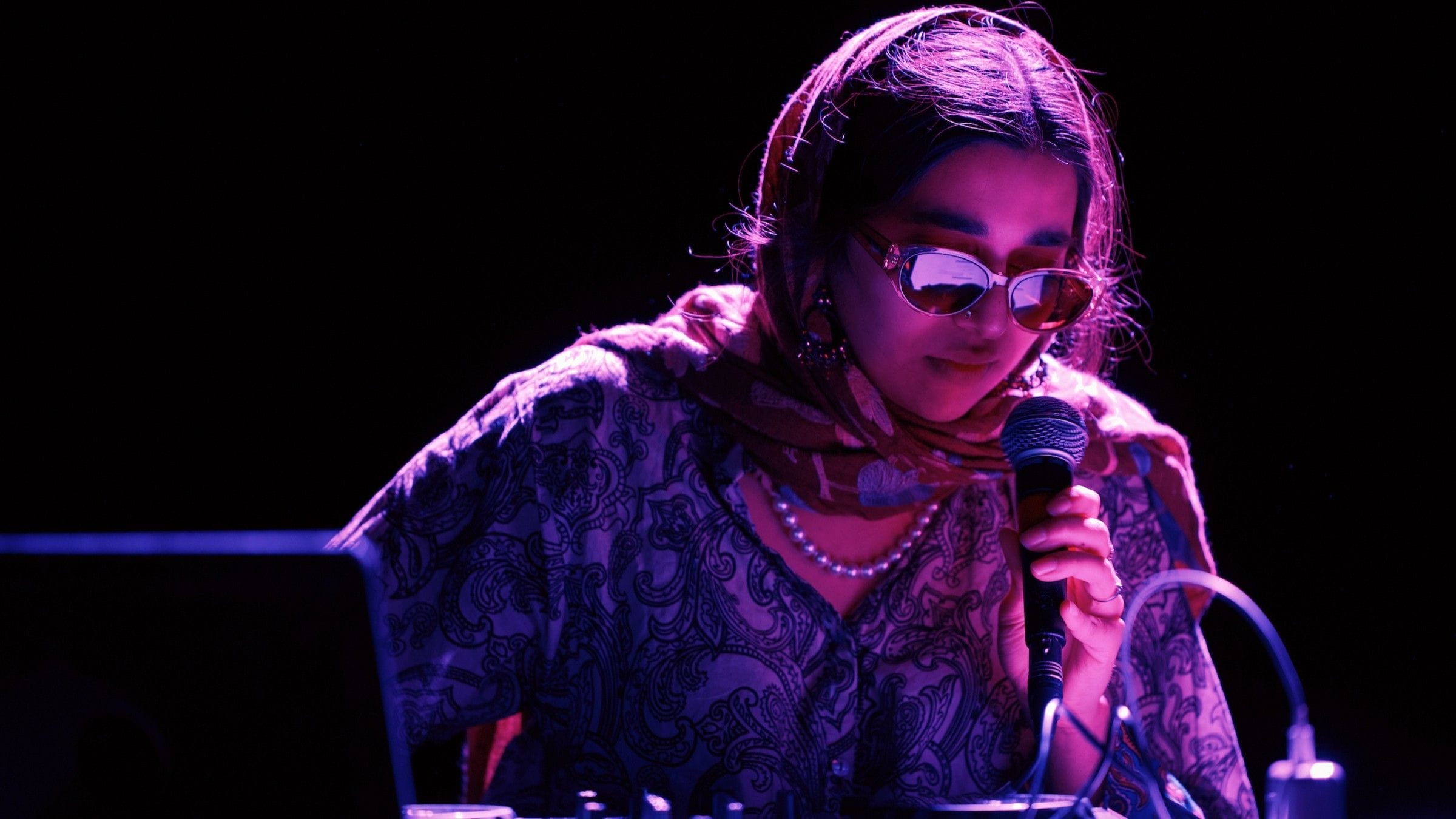 A person wearing sunglasses and a patterned scarf holds a microphone whilst operating audio equipment under purple lighting