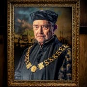 A framed portrait of an older man, reminiscent of A Man For All Seasons, wearing a black hat and elaborate dark clothing with a large gold chain, set against a dramatic background. The ornate frame features intricate decorative patterns.