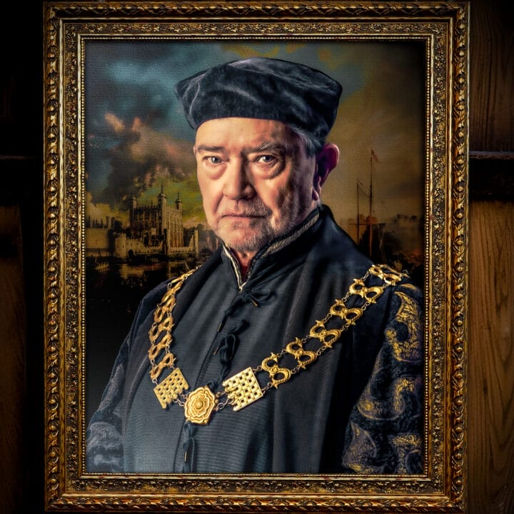 A framed portrait of an older man, reminiscent of A Man For All Seasons, wearing a black hat and elaborate dark clothing with a large gold chain, set against a dramatic background. The ornate frame features intricate decorative patterns.