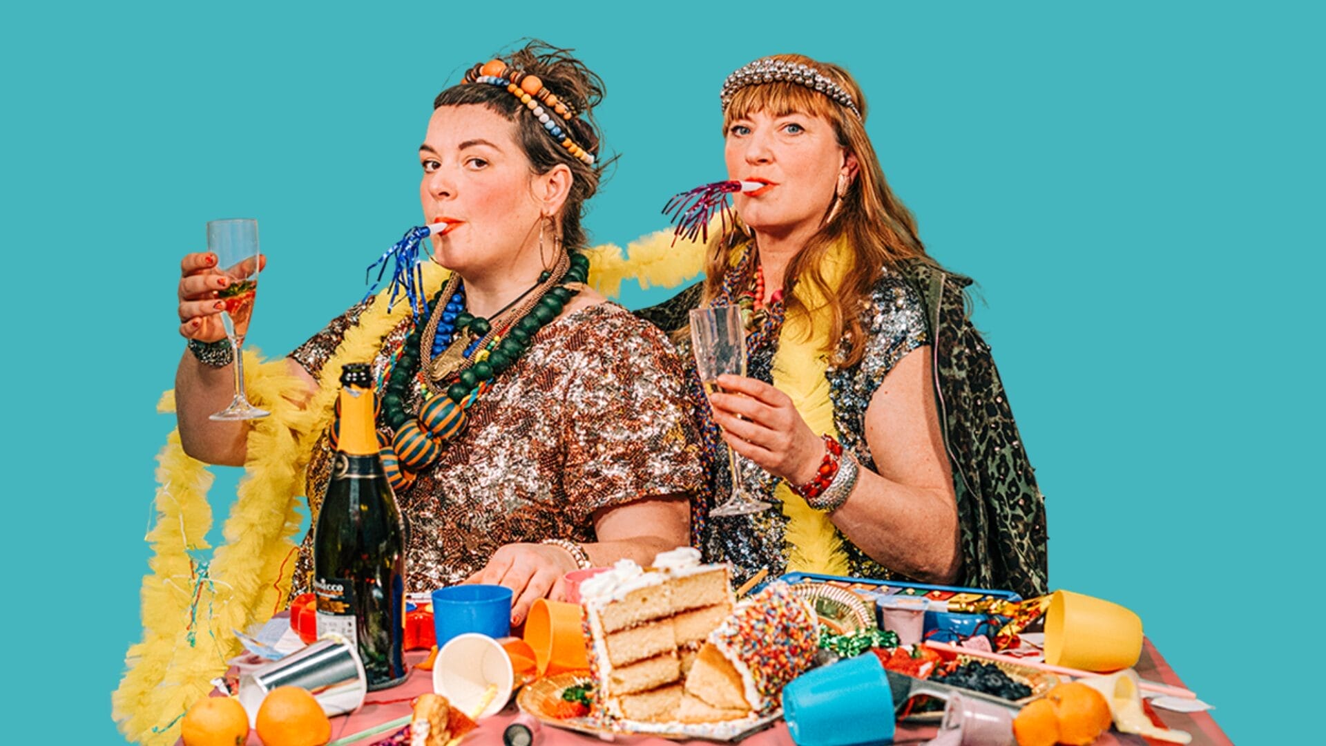 Two women dressed in festive, shiny clothing and bold accessories sit at a table with cake, confetti, fruit, and party items. Both are holding drinks with party blowers in their mouths against a plain turquoise background.