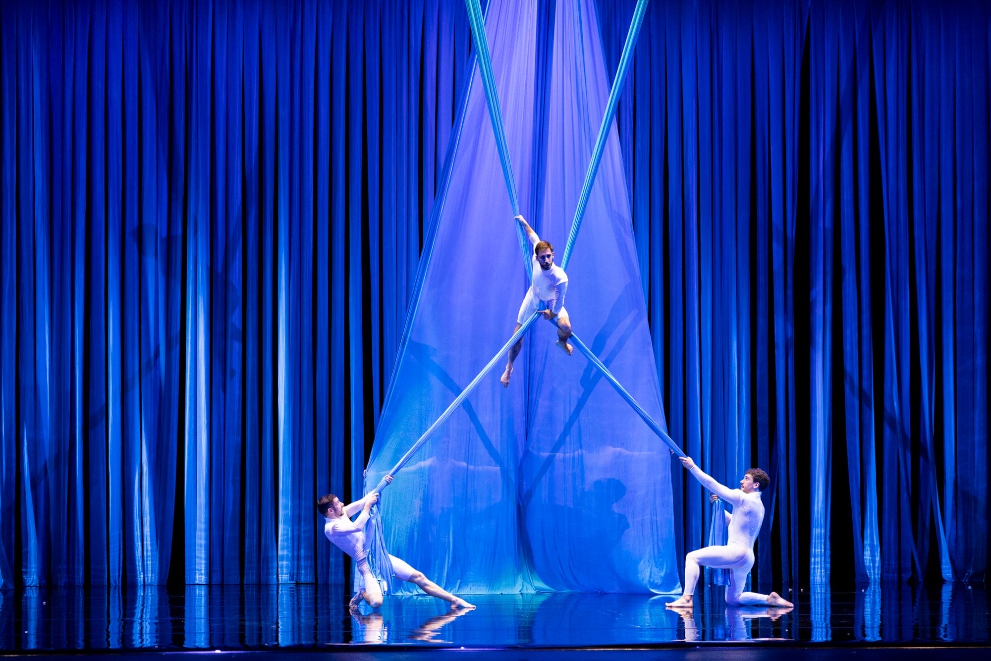 Three aerial performers in white bodysuits use blue silk fabrics for an acrobatic routine on stage, with blue curtains and dramatic lighting in the background.