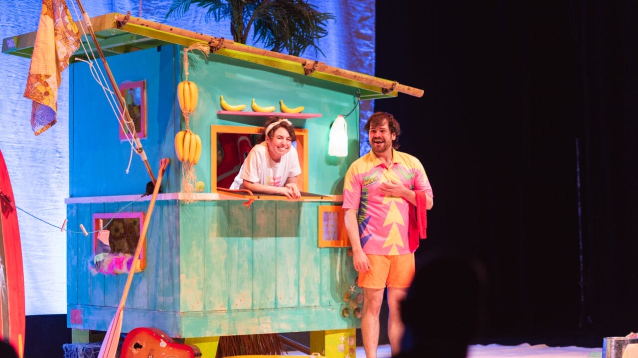 Two people stand by a colourful beach hut on stage; one leans from a window smiling, while the other—in a bright shirt with bananas—stands in front. A guitar, beach props, and touches of Christmas décor are visible nearby.