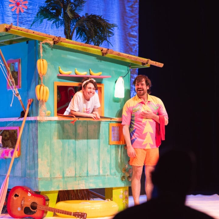 Two people stand by a colourful beach hut on stage; one leans from a window smiling, while the other—in a bright shirt with bananas—stands in front. A guitar, beach props, and touches of Christmas décor are visible nearby.