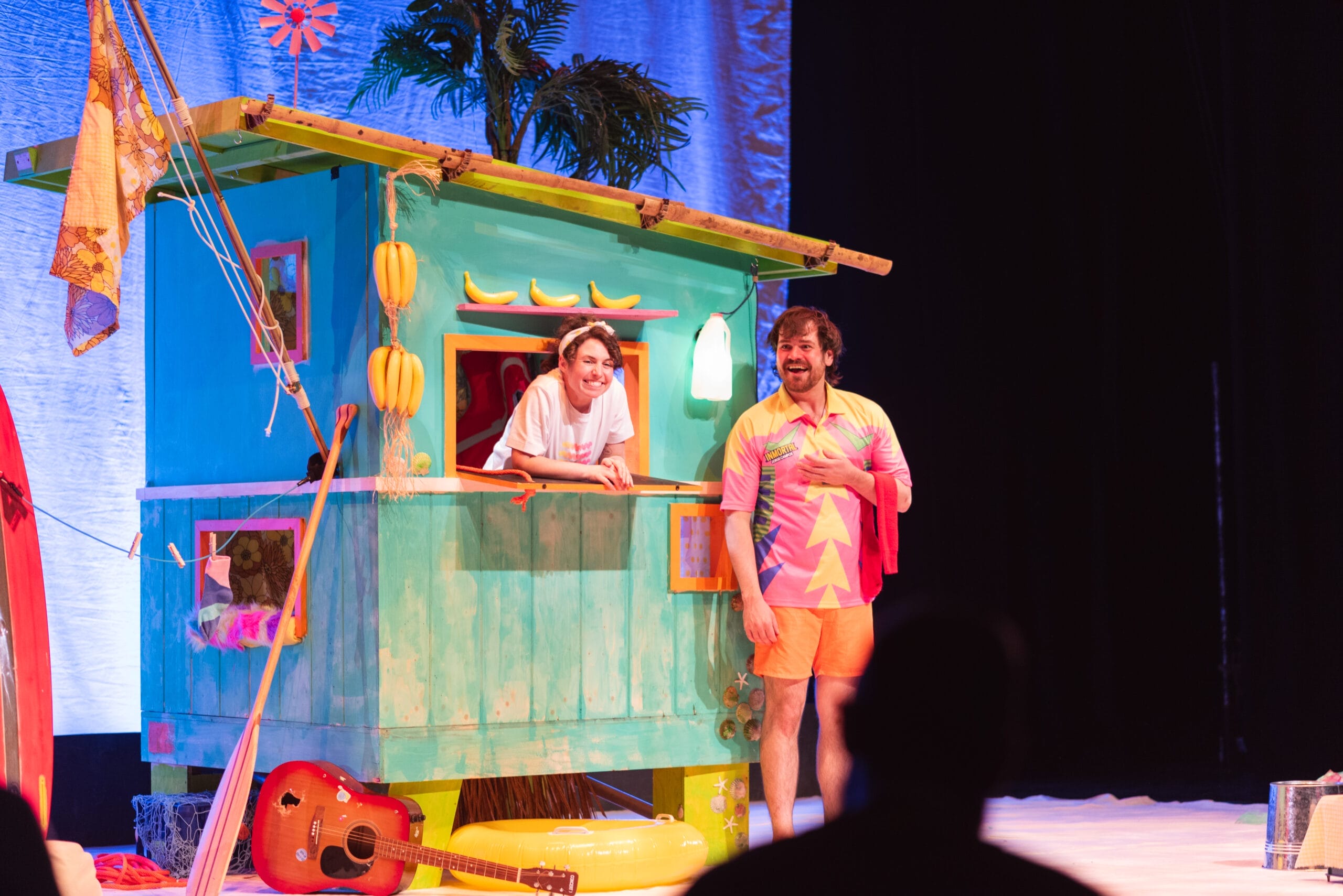 Two people stand by a colourful beach hut on stage; one leans from a window smiling, while the other—in a bright shirt with bananas—stands in front. A guitar, beach props, and touches of Christmas décor are visible nearby.