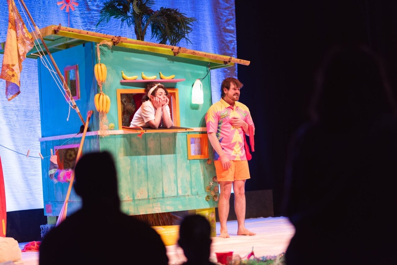 Two performers are on stage; one leans out of a colourful, banana-themed hut, while the other stands beside it in a pink and orange outfit. Silhouettes of audience members are visible in the foreground.