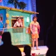 Two performers are on stage; one leans out of a colourful, banana-themed hut, while the other stands beside it in a pink and orange outfit. Silhouettes of audience members are visible in the foreground.