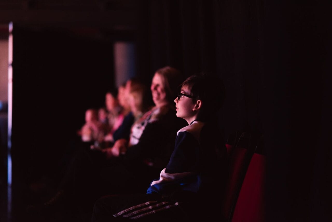 A boy wearing glasses sits in a dimly lit theatre, looking ahead, perhaps awaiting a Christmas performance. Several adults and other children sit in a row behind him, all facing the same direction against the dark background.