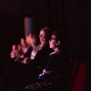 A boy wearing glasses sits in a dimly lit theatre, looking ahead, perhaps awaiting a Christmas performance. Several adults and other children sit in a row behind him, all facing the same direction against the dark background.