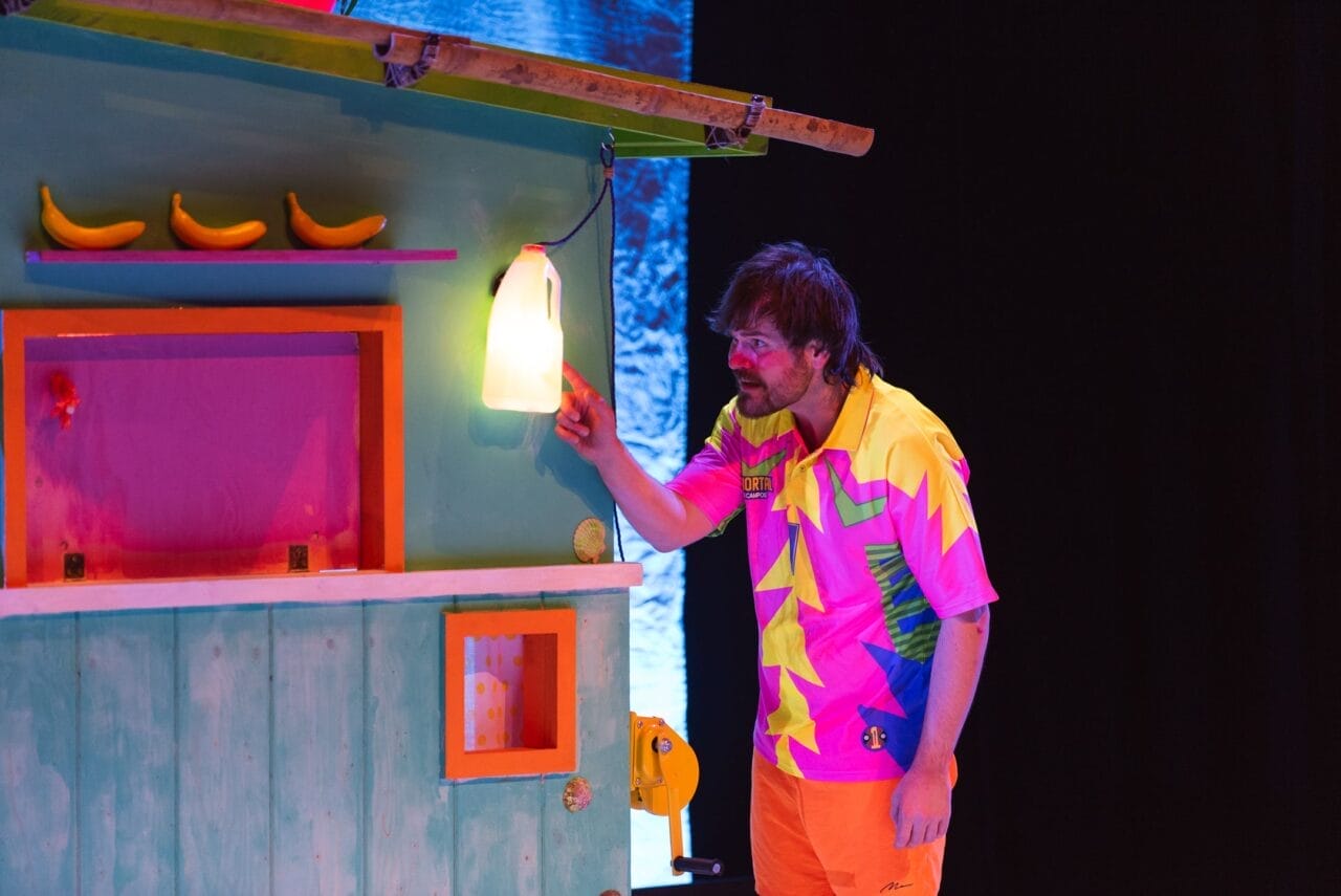 A man in a colourful, patterned outfit touches a glowing jug attached to a brightly painted structure with bananas and a small window, all set against a dark, Christmas-lit background.