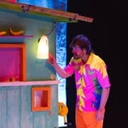 A man in a colourful, patterned outfit touches a glowing jug attached to a brightly painted structure with bananas and a small window, all set against a dark, Christmas-lit background.