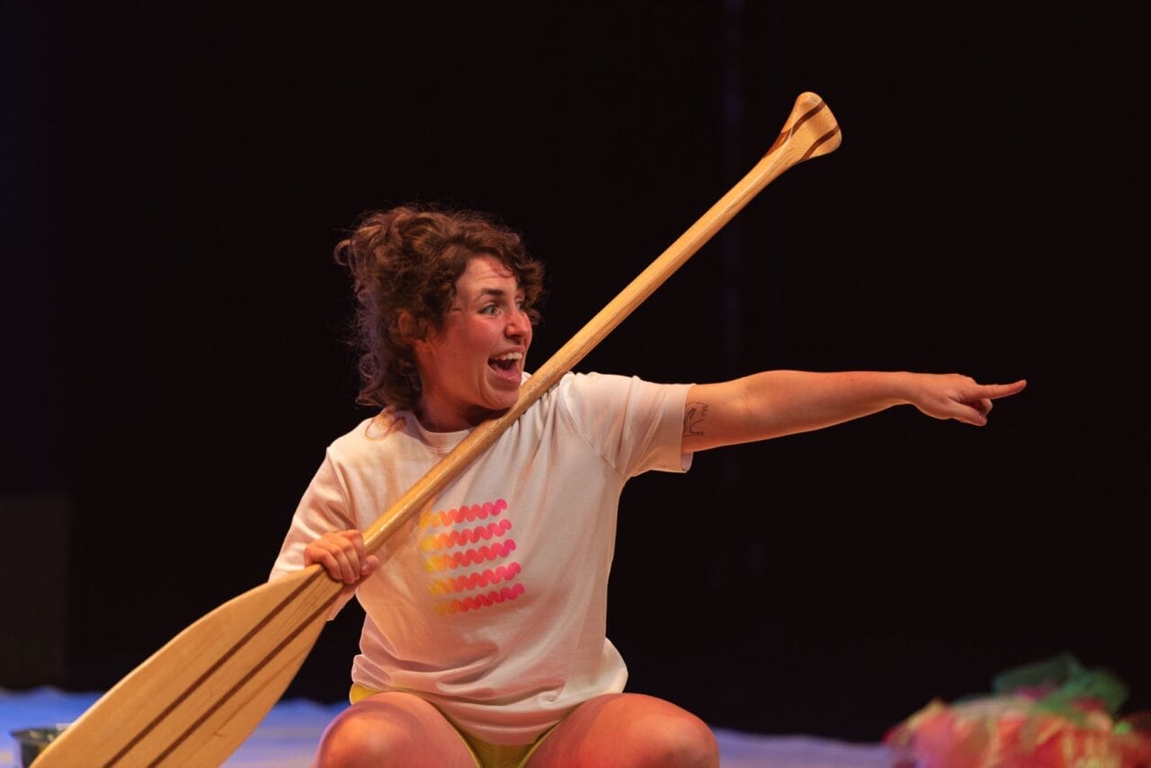 A person with curly hair holds a wooden bat and points excitedly to the right. Wearing a white T-shirt with a colourful dot pattern and shorts, they sit against a dark background, looking ready for a game—or perhaps some Christmas fun with bananas.