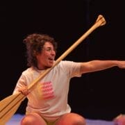 A person with curly hair holds a wooden bat and points excitedly to the right. Wearing a white T-shirt with a colourful dot pattern and shorts, they sit against a dark background, looking ready for a game—or perhaps some Christmas fun with bananas.