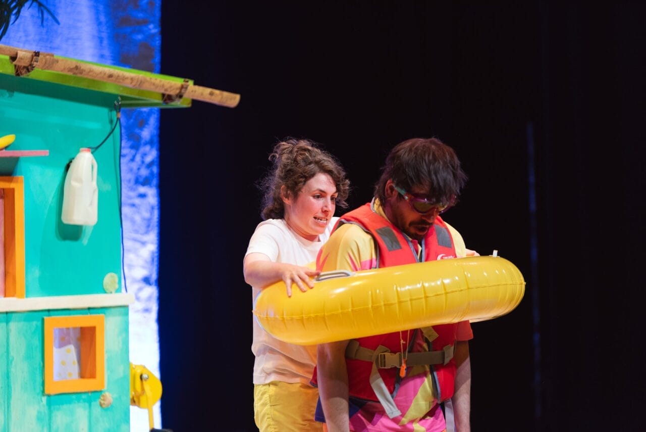 A woman stands behind a man wearing a life jacket and a yellow inflatable swim ring shaped like bananas. The woman appears to be assisting him next to a colourful, green and yellow structure with a dark background.