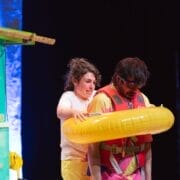 A woman stands behind a man wearing a life jacket and a yellow inflatable swim ring shaped like bananas. The woman appears to be assisting him next to a colourful, green and yellow structure with a dark background.