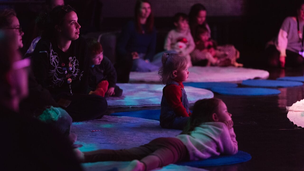 Children and adults sit and lie on mats and rugs on a dark floor, watching something off-camera. The environment is dimly lit with purple and blue lighting, evoking a cosy Christmas atmosphere.