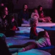 Children and adults sit and lie on mats and rugs on a dark floor, watching something off-camera. The environment is dimly lit with purple and blue lighting, evoking a cosy Christmas atmosphere.