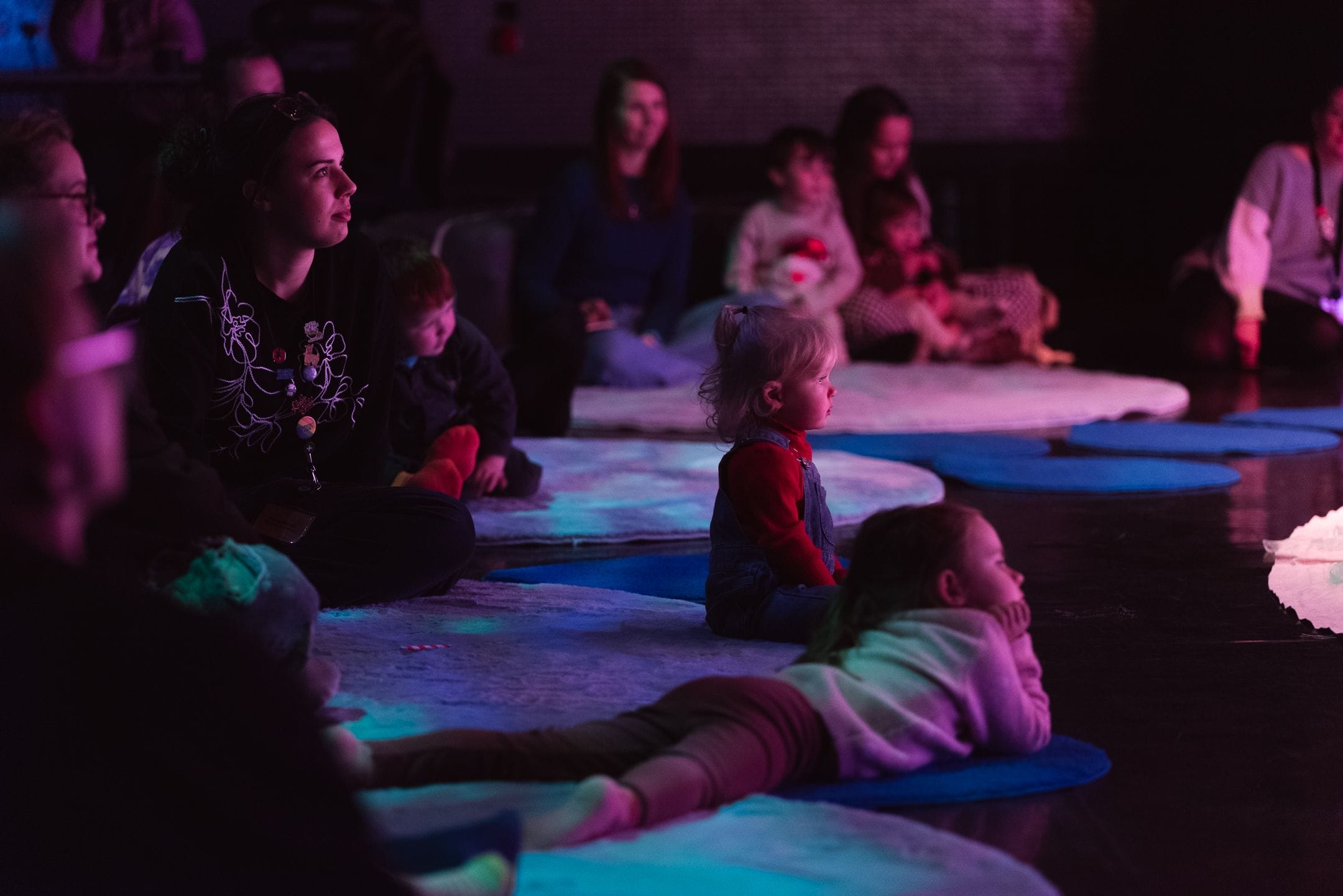 Children and adults sit and lie on mats and rugs on a dark floor, watching something off-camera. The environment is dimly lit with purple and blue lighting, evoking a cosy Christmas atmosphere.