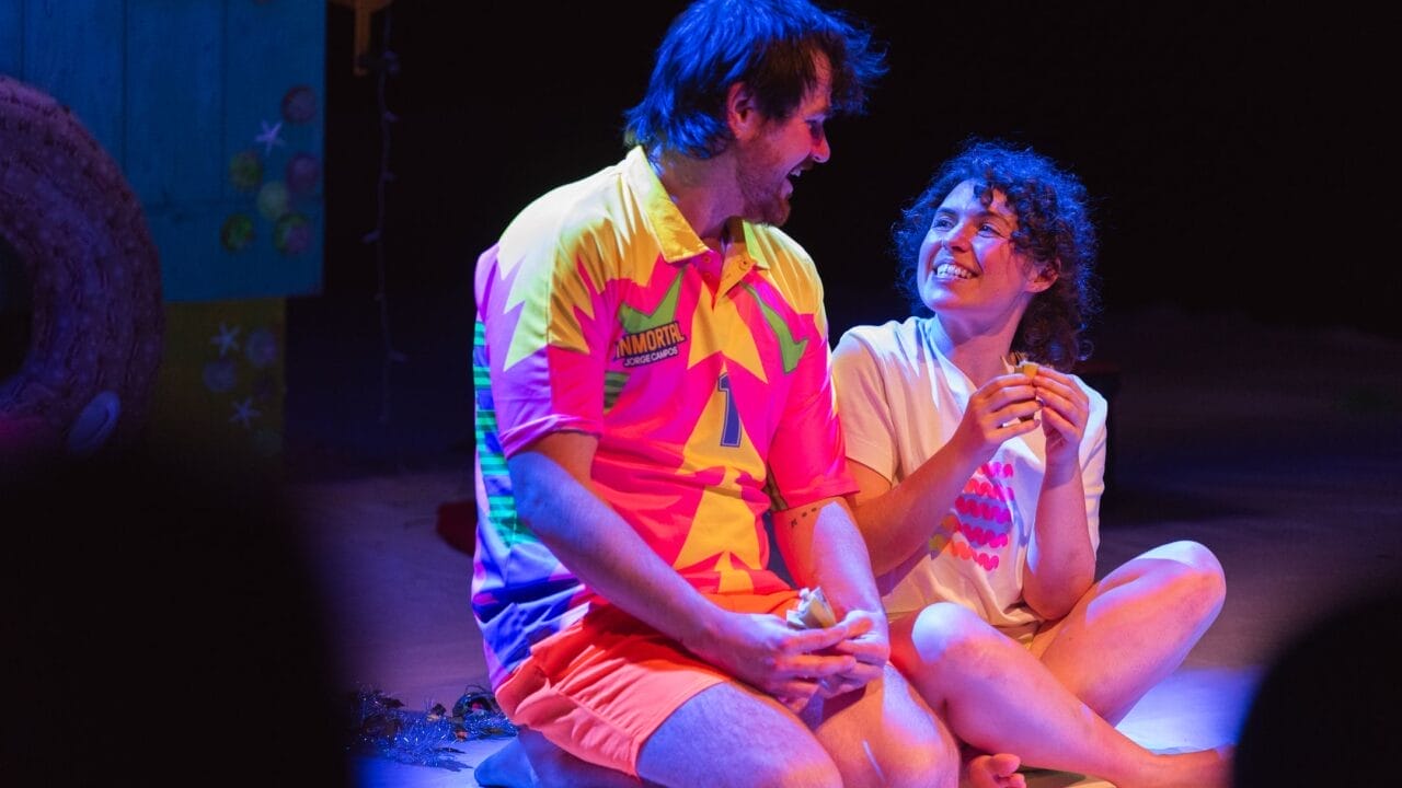 A man in a colourful shirt and orange shorts sits cross-legged onstage next to a smiling woman in a white shirt. Amidst stage props and a painted set, they chat animatedly—perhaps planning for Christmas or sharing laughs over bananas.