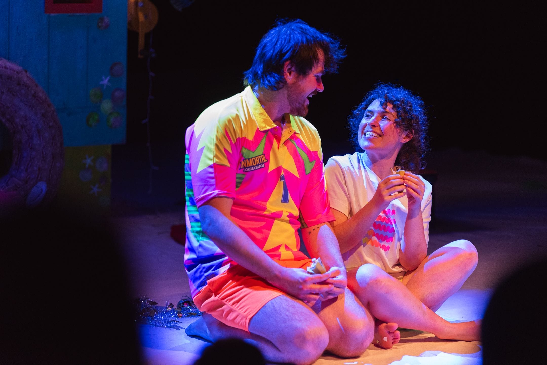 A man in a colourful shirt and orange shorts sits cross-legged onstage next to a smiling woman in a white shirt. Amidst stage props and a painted set, they chat animatedly—perhaps planning for Christmas or sharing laughs over bananas.