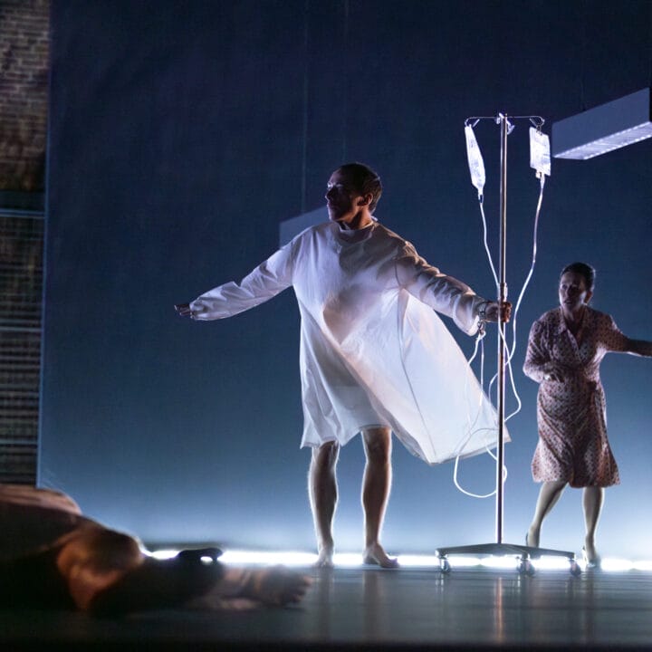 A person in a hospital gown stands with arms outstretched next to a drip stand, while a woman in a dress stands behind them.