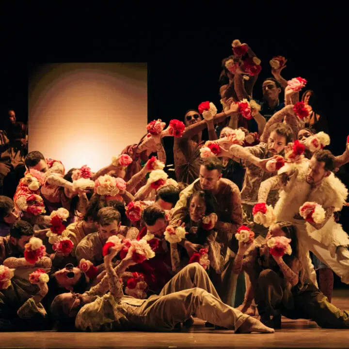 A group of dancers on stage pose closely together, holding red and white flowers. They wear earth-toned costumes, and warm lighting highlights their expressive gestures against a dark background.