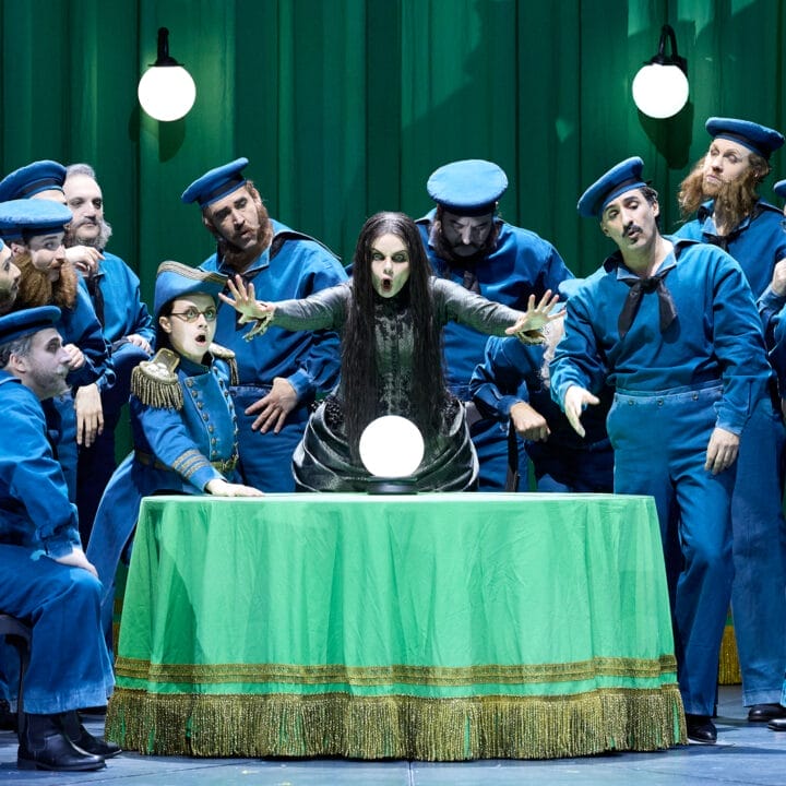 A woman with long dark hair sits at a green-covered table, her hands hovering over a glowing orb. She is surrounded by men in blue sailor uniforms against a green curtain backdrop.