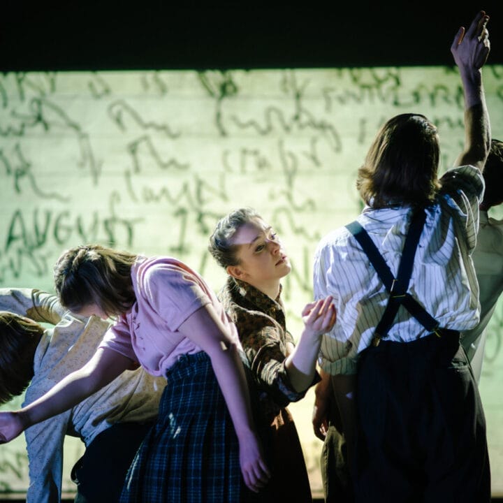 Five people stand closely together, leaning and reaching with arms outstretched, in front of a large screen displaying handwritten text. The lighting highlights the performers and their expressive poses.