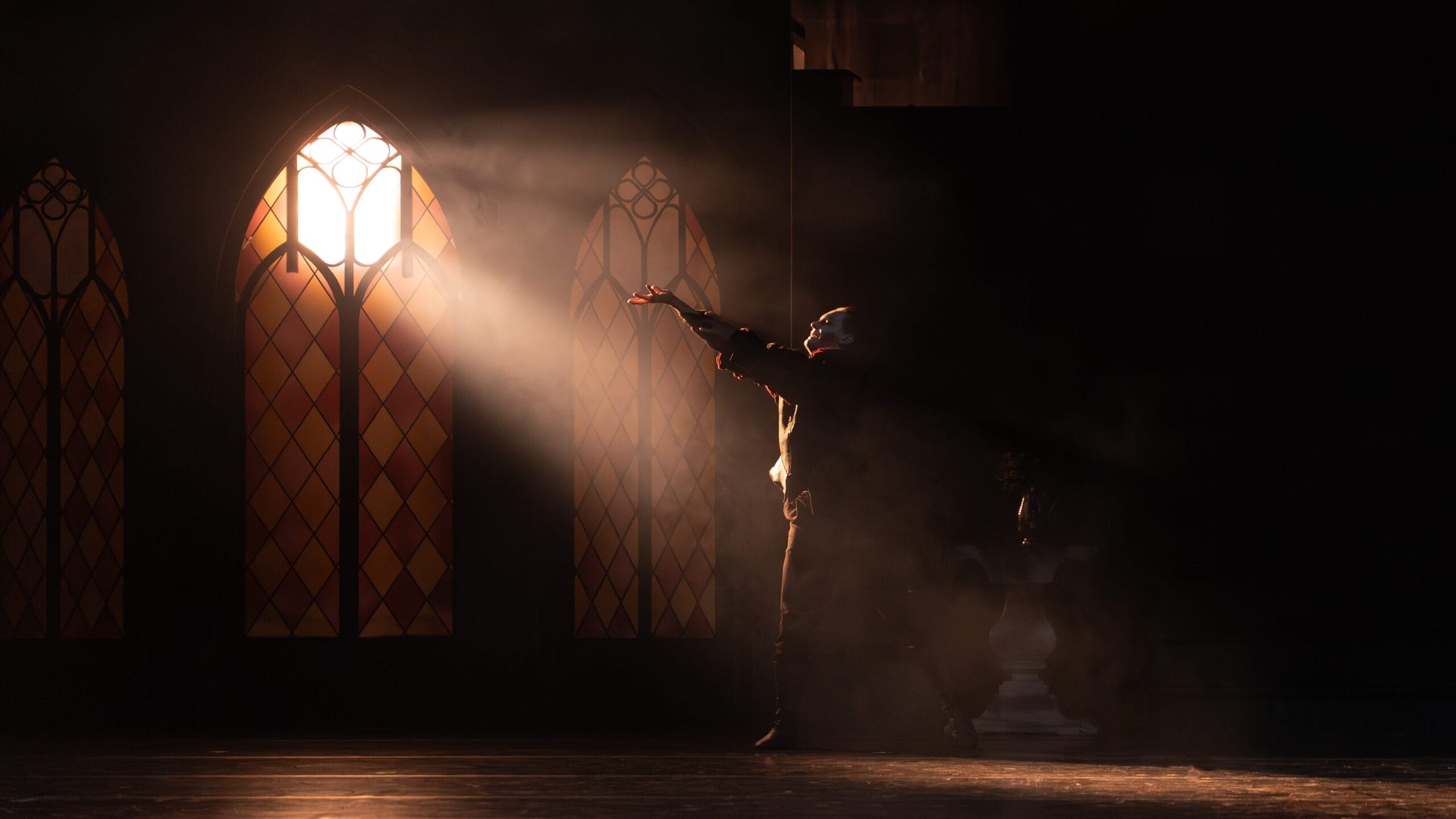 A person wearing dark clothing and a mask stands on a dimly lit stage, reaching towards a beam of light coming through a tall, arched stained-glass window. Dust is visible in the light.