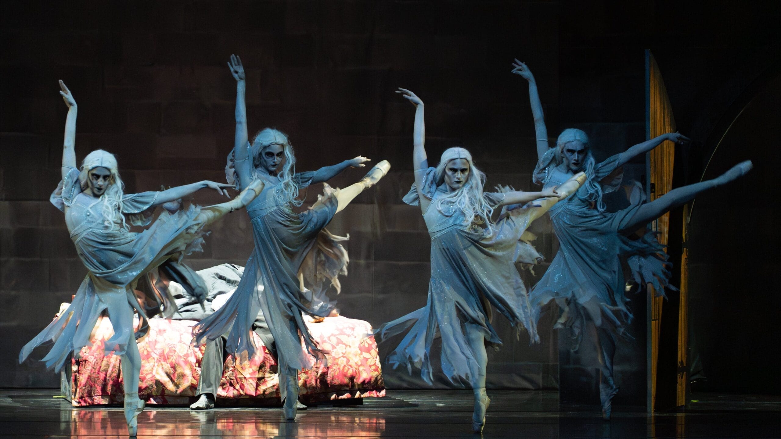 Four dancers in tattered, ghost-like costumes with white make-up perform synchronised ballet poses on stage