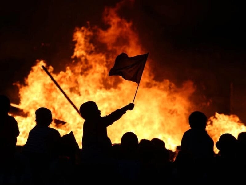 Silhouettes of children are seen in front of a large fire at night. One child holds a flag, and another pole is visible. The flames create a bright, dramatic background.