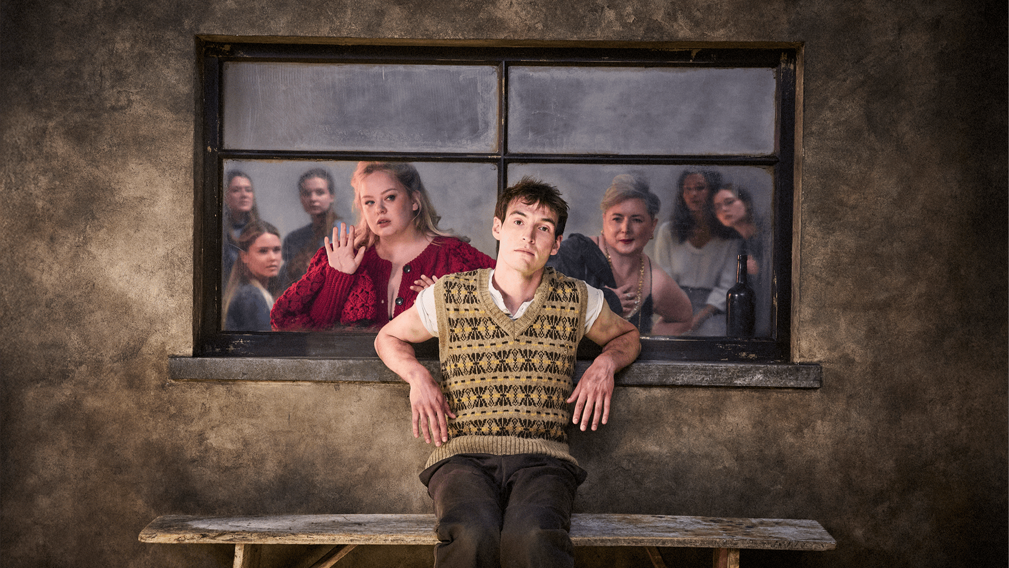 A young man in a patterned waistcoat sits on a bench in front of a concrete wall. Behind him, a group of women, recalling J.M. Synge's *Playboy of the Western World*, look through a window with concerned or serious expressions.