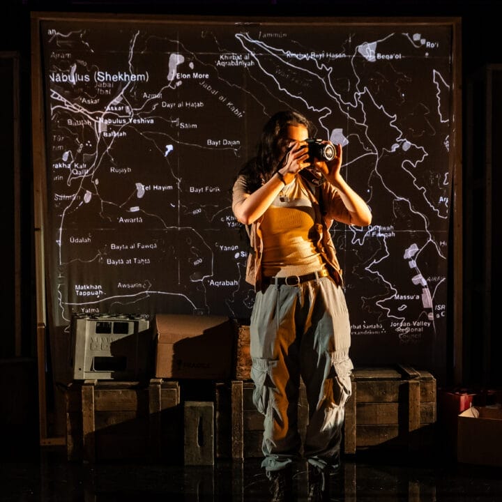 A person stands on stage holding a camera, with a projected map of Nablus (Shekhem) and nearby locations displayed behind them.