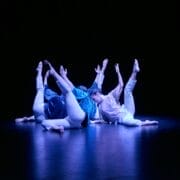 Four dancers dressed in blue and white costumes lie on a dark stage, raising their arms and legs upward in unison under a spotlight, showcasing dance innovations that push the boundaries of movement on the Dance Horizons stage.
