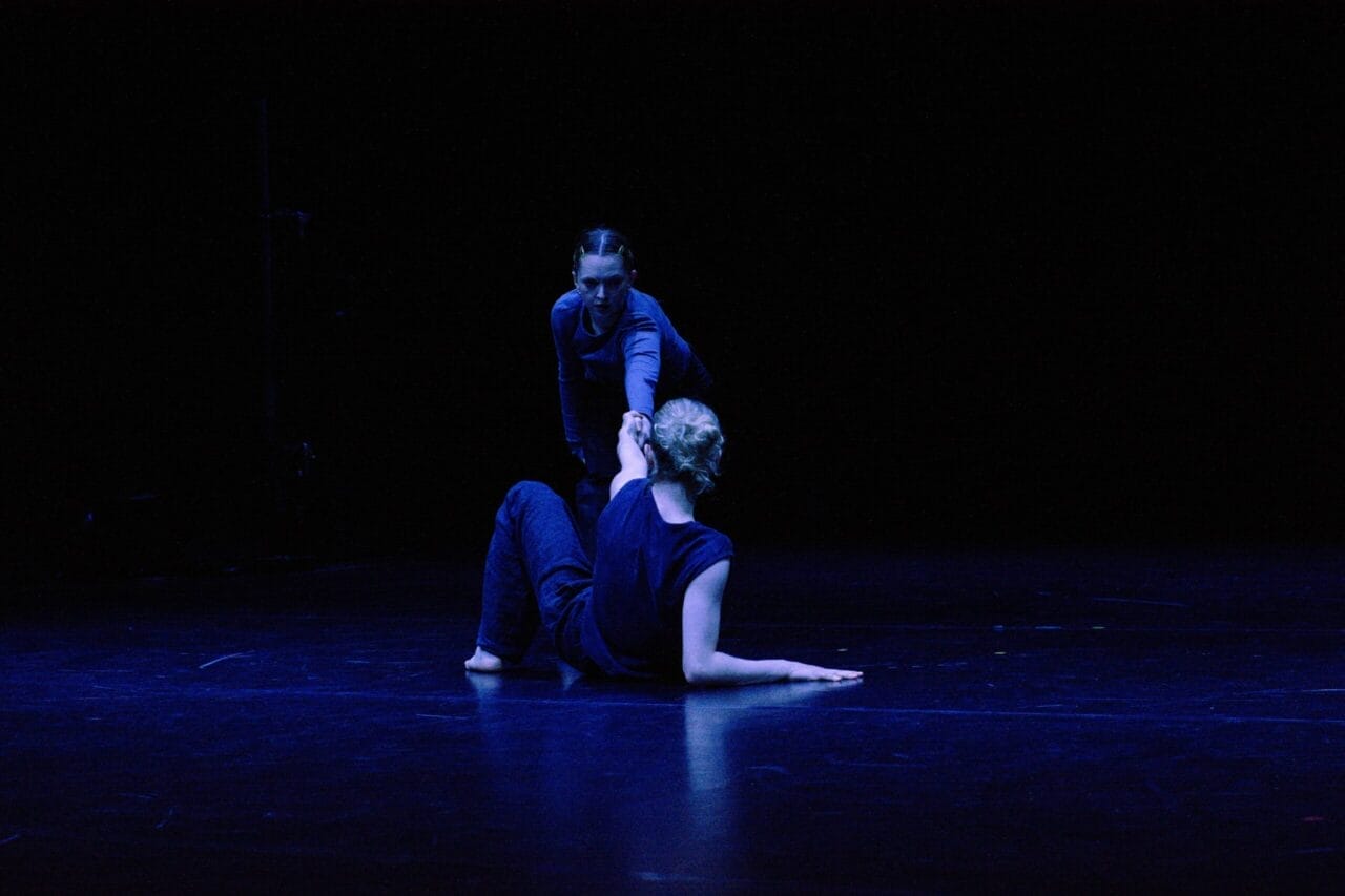 Two people perform contemporary dance on a dimly lit stage. As blue lighting highlights the scene, their expressive movements capture the spirit of Dance Innovations against a dark background.