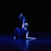 Two people perform contemporary dance on a dimly lit stage. As blue lighting highlights the scene, their expressive movements capture the spirit of Dance Innovations against a dark background.