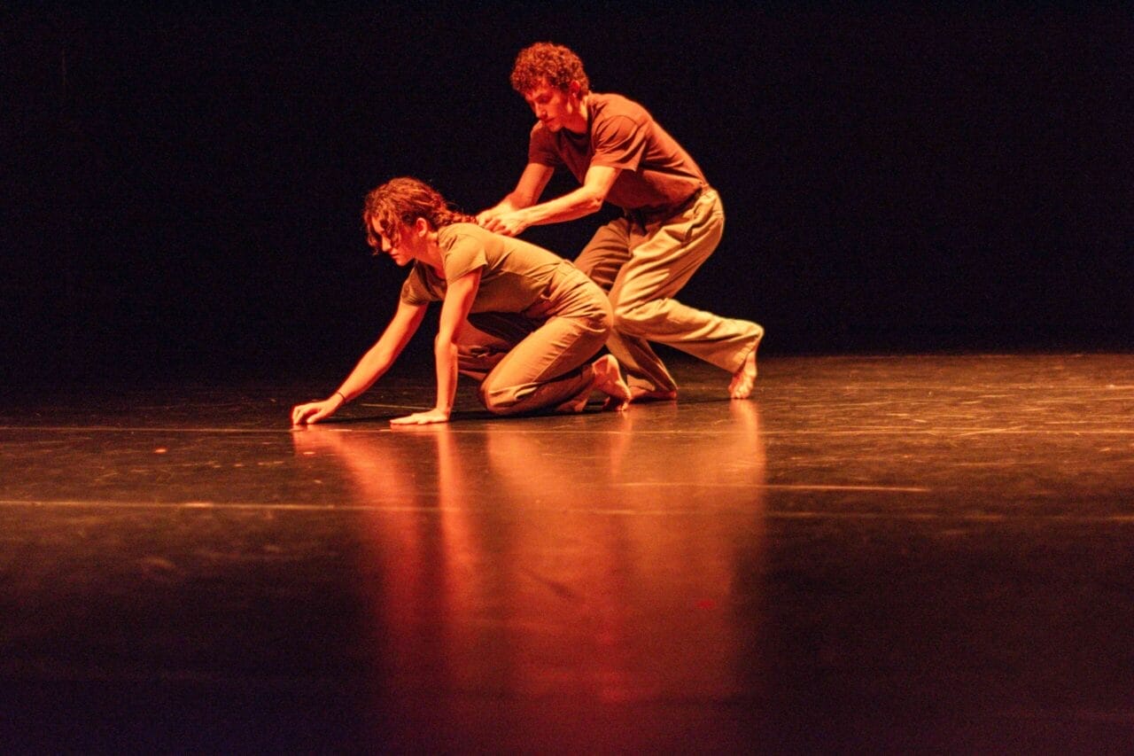 Two people on stage perform a modern dance from Dance Horizons. One kneels on all fours with a hand reaching forward, whilst the other crouches behind, placing hands gently on their back. The stage is dark with orange lighting.