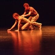 Two people on stage perform a modern dance from Dance Horizons. One kneels on all fours with a hand reaching forward, whilst the other crouches behind, placing hands gently on their back. The stage is dark with orange lighting.